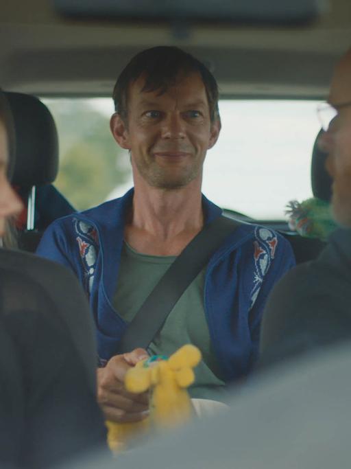 Filmstill aus "Whatever Happens Next" von Regisseur Julian Pörksen: die Hauptfigur Paul sitzt auf dem Rücksitz eines Autos, auf dem Fahrer- und Beifahrersitz ist ein Paar zu sehen