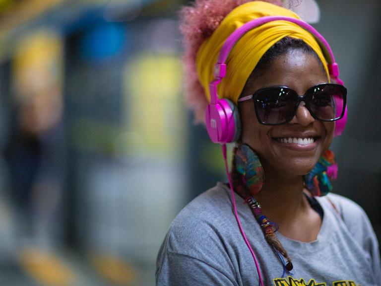 Eine Frau steht vor einem Zug und hört sichtlich glücklich Musik über Kopfhörer.