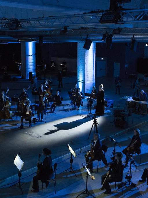 Ein Orchester sitzt in einer blau ausgeleuchteten Industriehalle. In der Mitte stehen Dirigent und Sängerin auf einer Metallplattform.