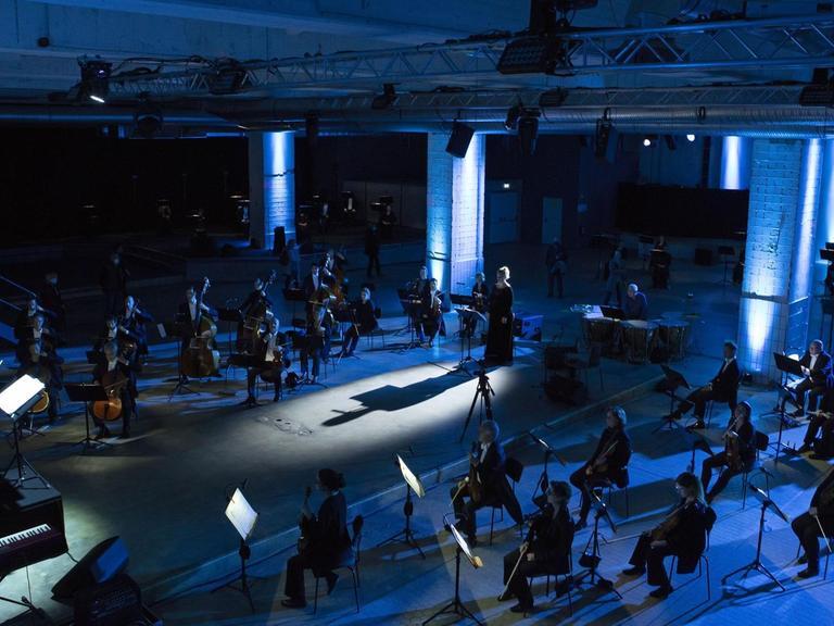 Ein Orchester sitzt in einer blau ausgeleuchteten Industriehalle. In der Mitte stehen Dirigent und Sängerin auf einer Metallplattform.