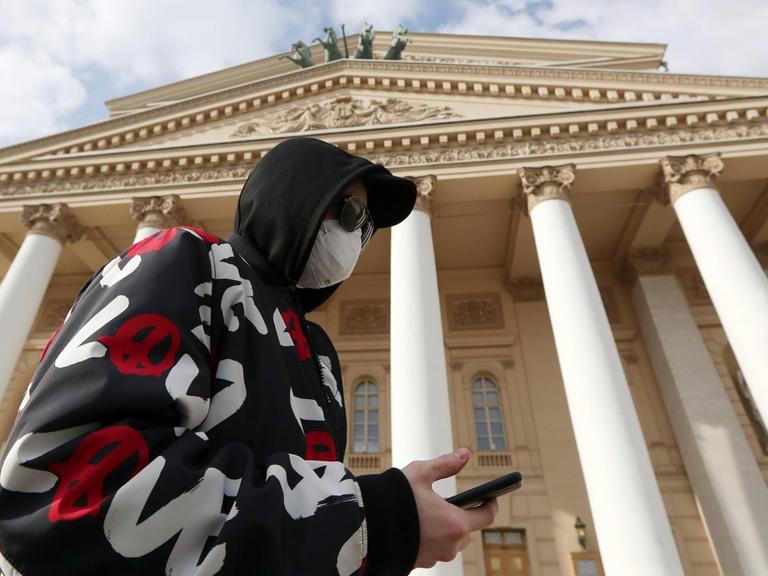 Ein Mann mit Smartphone, Mundschutz und Kapuzenpullover vor dem Bolshoi Theater in Moskau.
