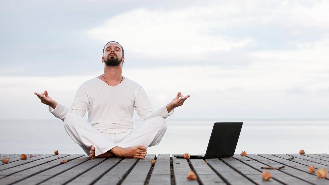 Ein weißgekleideter, meditierender Mann mit Laptop am Strand.
