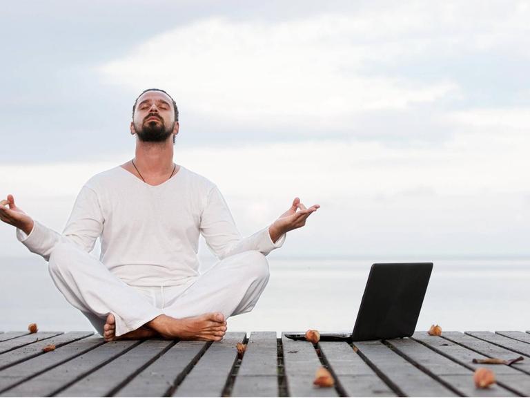 Ein weißgekleideter, meditierender Mann mit Laptop am Strand.