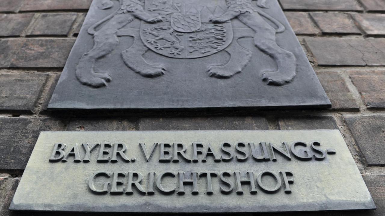 Das Schild hängt an einer Außenwand aus Backsteinen, darüber ein weiteres Schild mit dem bayerischen Wappen. Das Schild hängt an einer Außenwand aus Backsteinen, darüber ein weiteres Schild mit dem bayerischen Wappen.