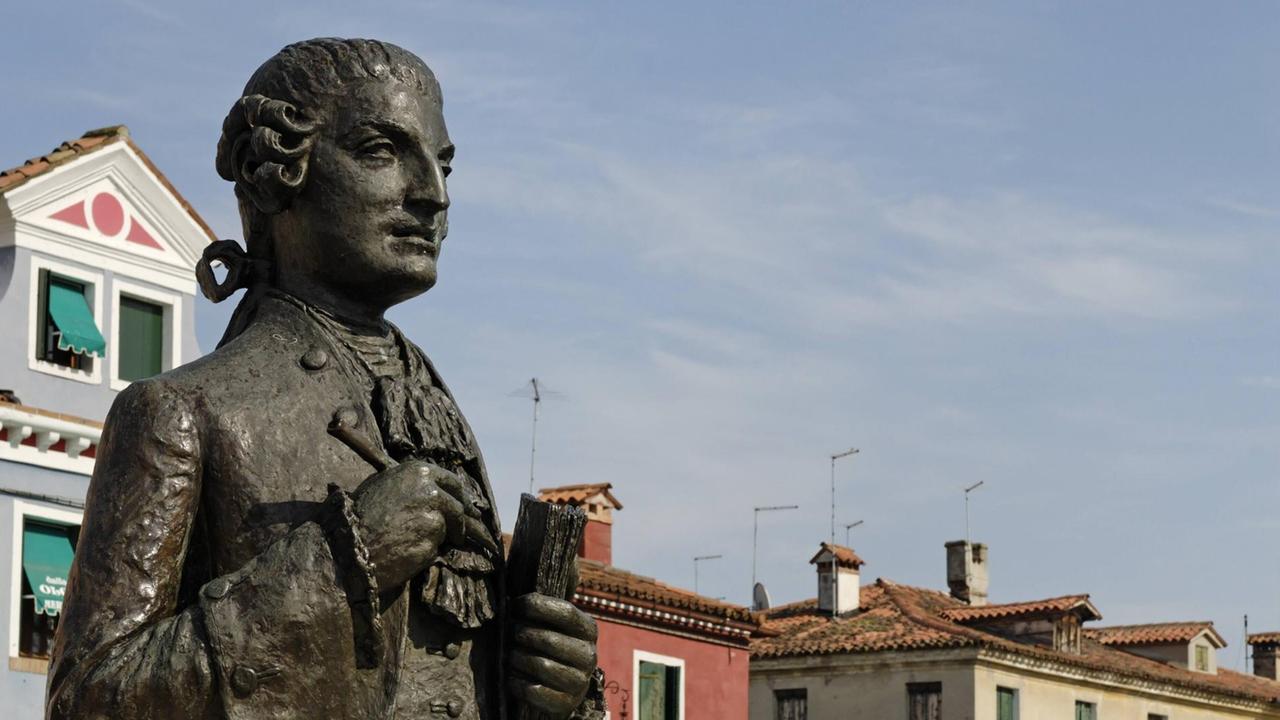 Die Statue von Baldassare Galuppi, die vor den bunten Häusern von Burano steht, hält einen Stift und ein Buch in den Händen.