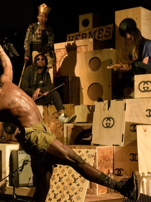 Bühnenszene mit Schauspielern vor und auf einem Berg aus Holzkisten, die die Schriftzüge von Luxusmarken tragen.