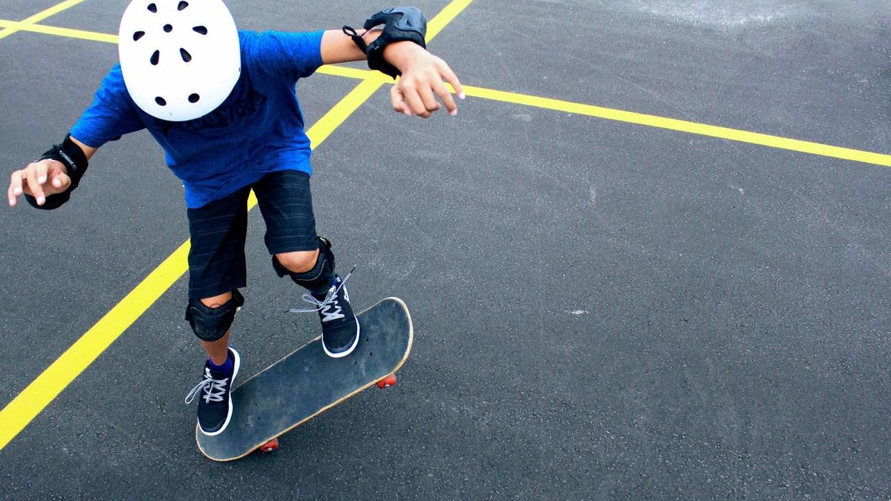 Ein Kind mit Helm und Schonern übt auf einer asphaltierten Fläche mit gelben Gitternetzlinien einen Trick auf dem Skateboard.