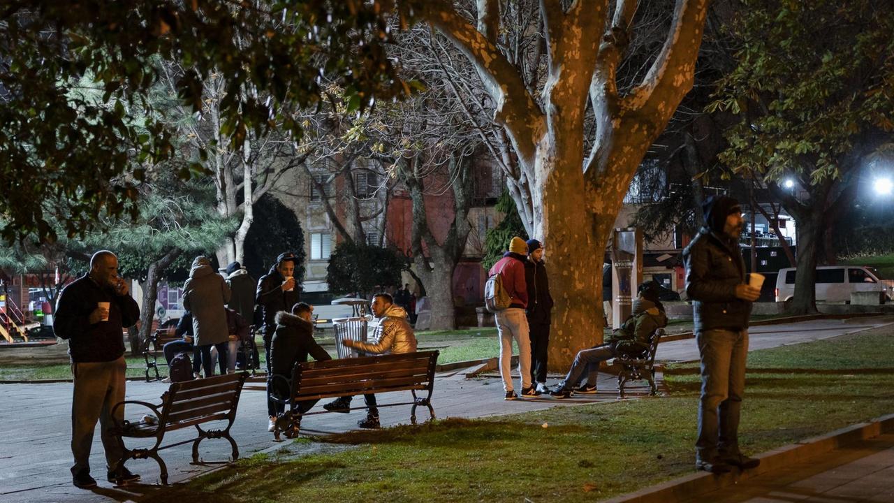 Mehrere Männer im Anorak stehen oder sitzen im Halbdunkel eines Parks und essen und trinken.