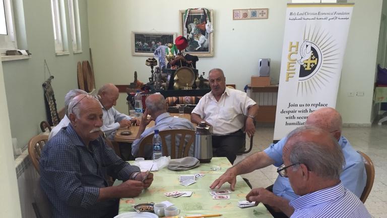 Senioren beim Kartenspiel im Sankt-Nicholas-Altenheim in Beit Jala. Im Hintergrund ist ein Plakat der 'Holy Land Christian Ecumenical Foundation' zu sehen.