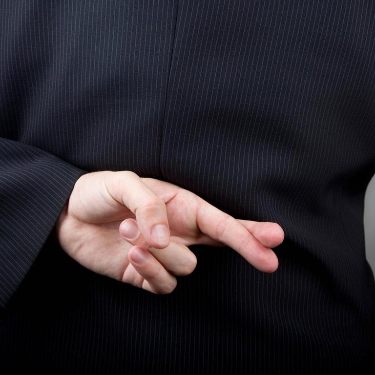 Ein Mann im Anzug hält seine Hand mit gekreuzten Fingern hinter den Rücken