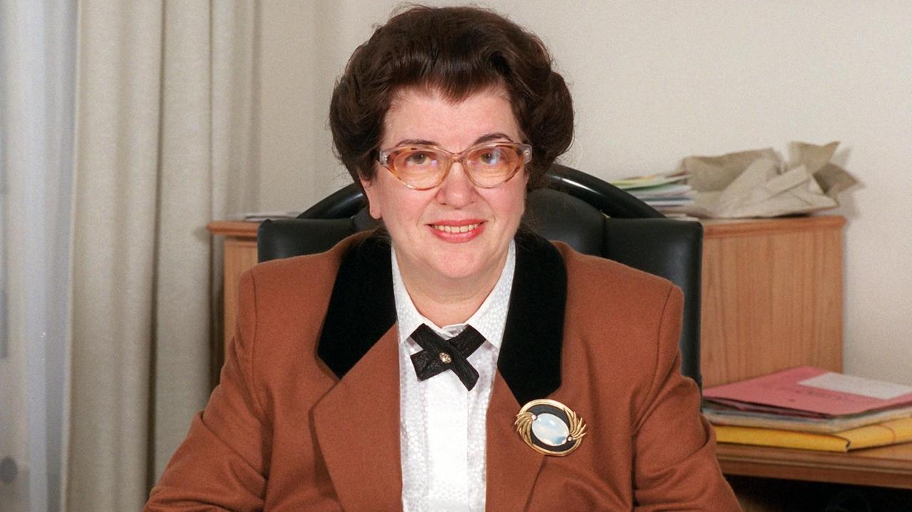 Lore Peschel-Gutzeit trägt einen braunen Blazer und sitzt an einem Schreibtisch. Im Hintergrund liegen Akten auf einem Tisch.