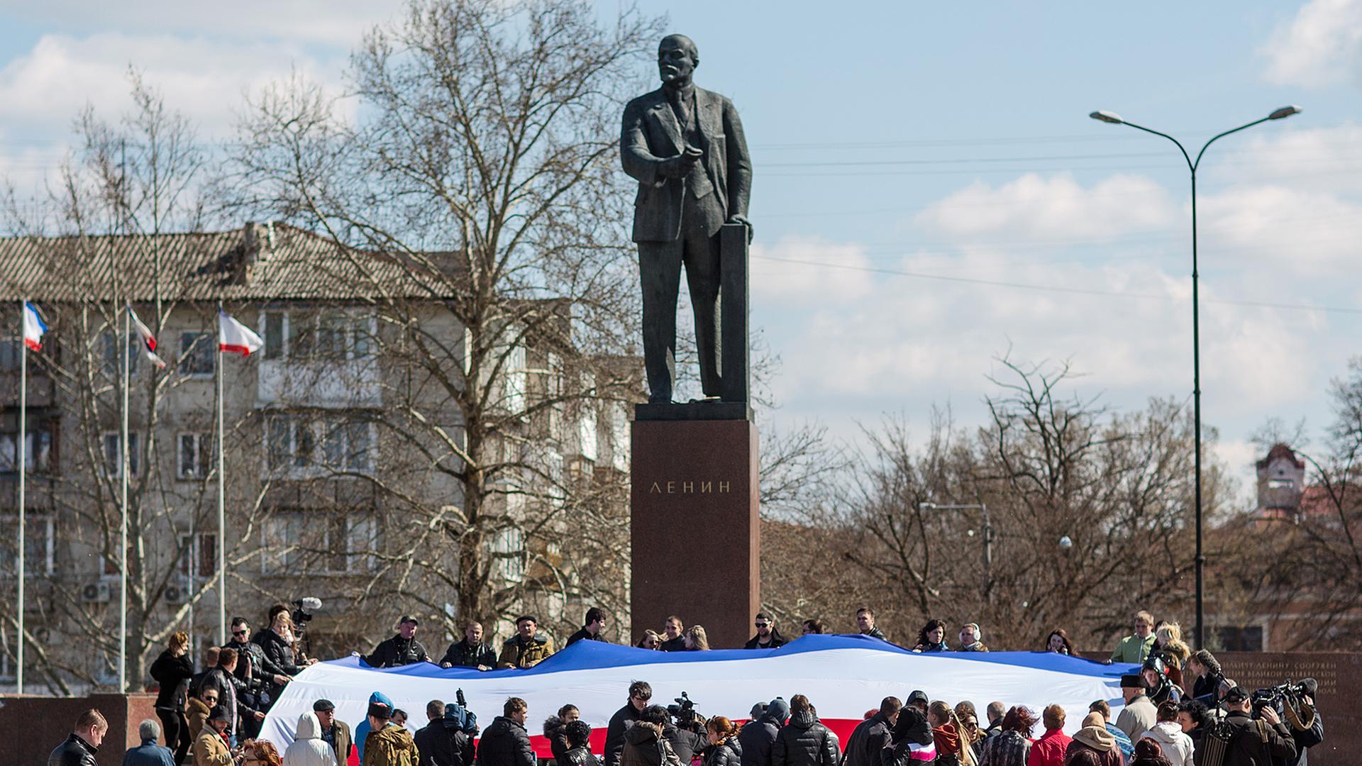 Dutzende Menschen halten am 18.03.2014 in Simferopol (Ukraine) anlässlich der Rede von Russlands Präsidenten Putin eine große russische Flagge vor dem Lenin-Denkmal in Händen.