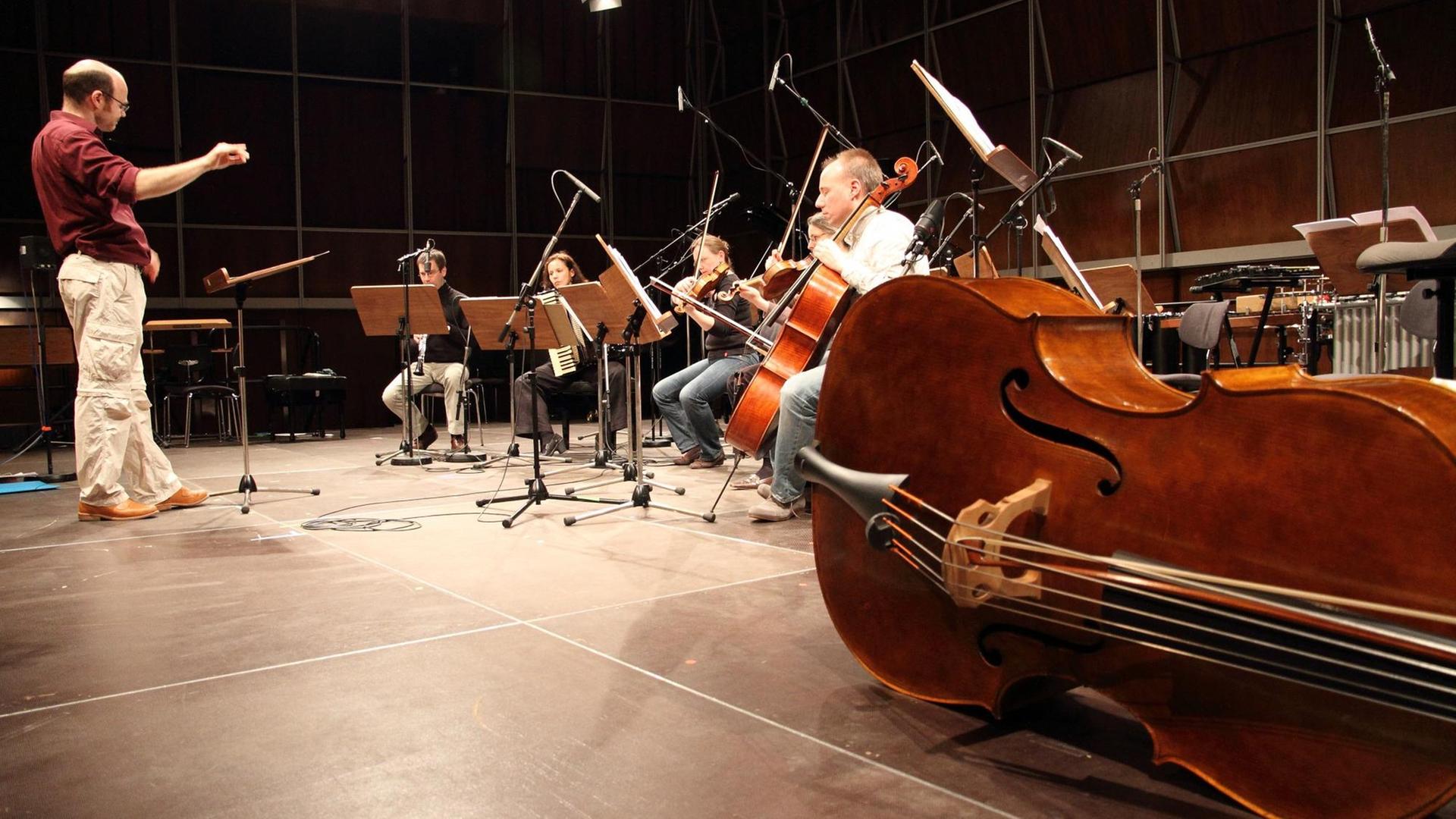 Fünf Musik stizen mit Instrumenten vor einem Dirigenten auf einer Bühne. Rechts von Ihnen liegt ein Kontrabass auf dem Boden. Fünf Musik stizen mit Instrumenten vor einem Dirigenten auf einer Bühne. Rechts von Ihnen liegt ein Kontrabass auf dem Boden.