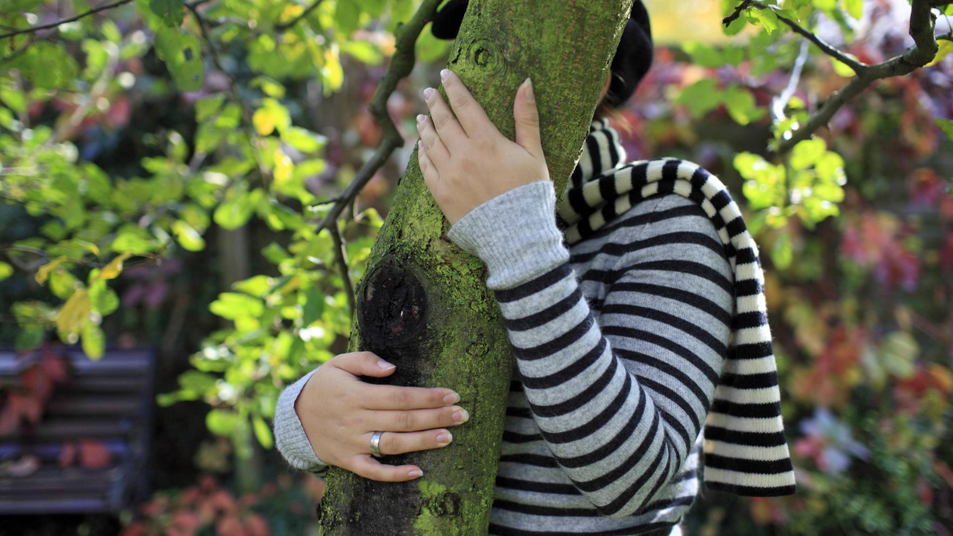 Eine junge Frau versteckt sich hinter einem Baumstamm.