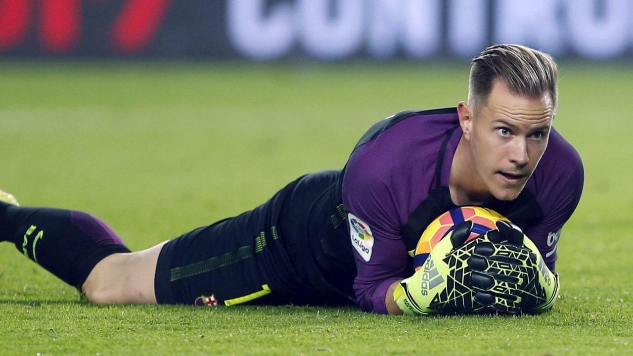 Ter Stegen liegt im lila Trikot auf dem Rasen und hat den Ball unter sich begraben. Er blickt konzentriert nach vorne. Ter Stegen liegt im lila Trikot auf dem Rasen und hat den Ball unter sich begraben. Er blickt konzentriert nach vorne.