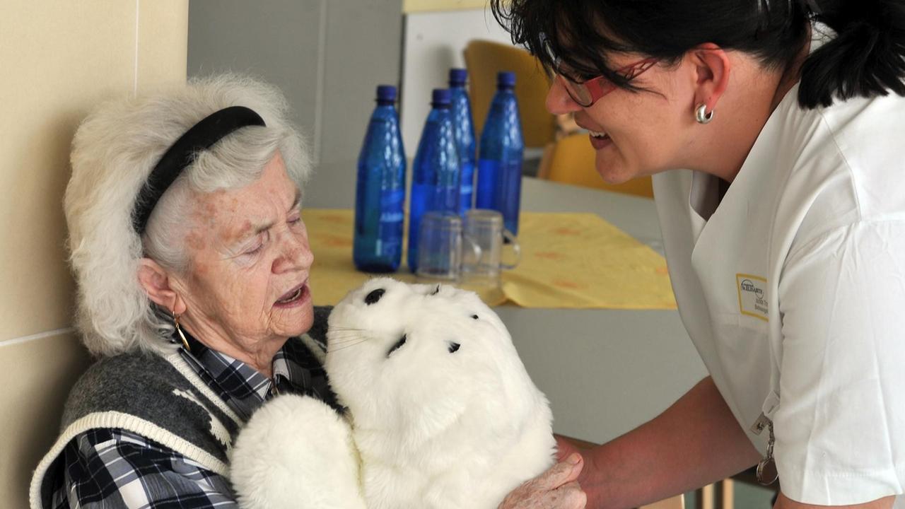 Eine Pflegekraft reicht der dementen Eleonore Mayer einen Roboter in einem Robbenfell, der sich bewegt und Geräusche macht