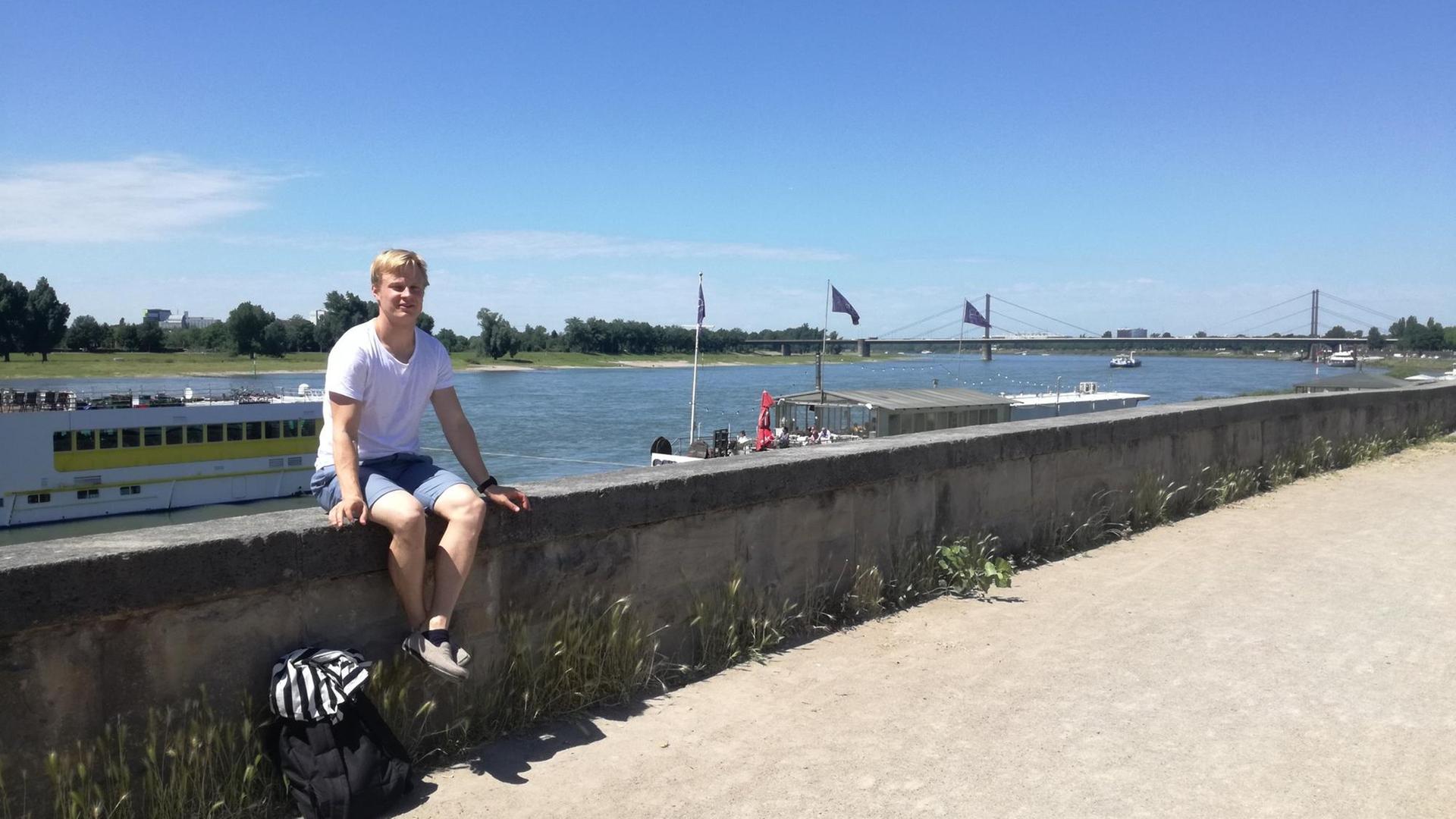 Ein junger Mann sitzt auf einer Mauer am Fluss Rhein. Hinter ihm sieht man Schiffe und eine Brücke.