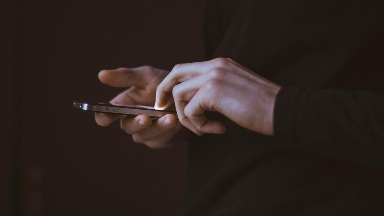 Zwei Hände tippen auf einem Smartphone vor schwarzem Hintergrund.