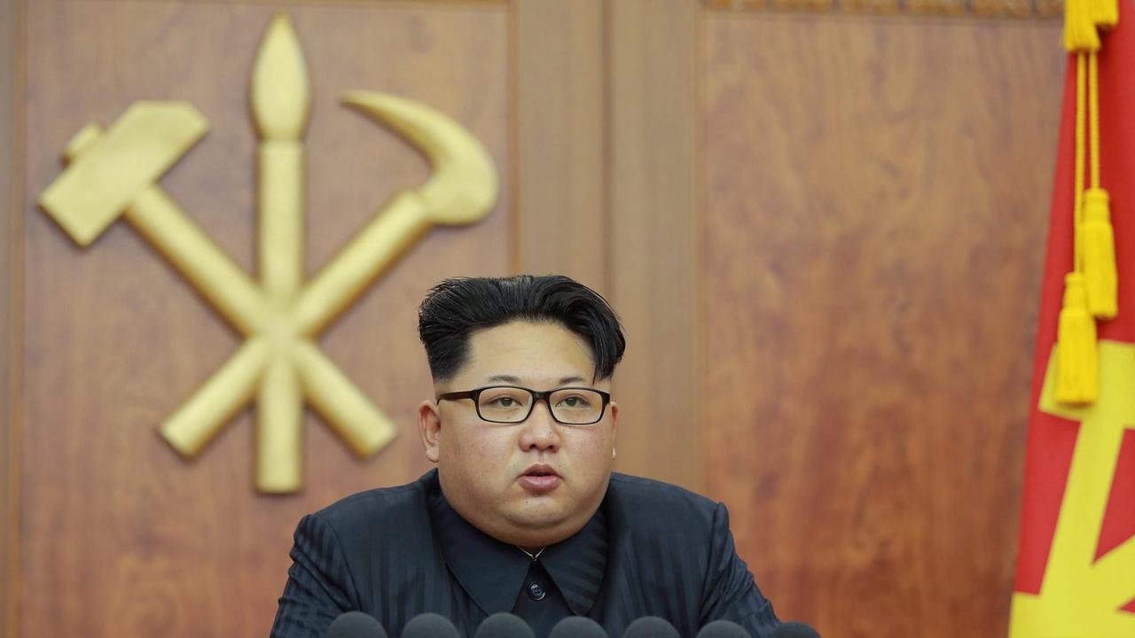 Zu sehen ist Nordkoreas Machthaber Kim Jong Un am Schreibtisch mit Brille, dahinter die Symbole Hammer, Sichel und Pinsel an einer Holzwand.