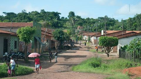 Eine Frau mit Sonnenschirm läuft auf der ungeteerten Straße eines Dorfes in Codó im Norden Brasiliens, rechts und links der Straße stehen kleine Häuser.