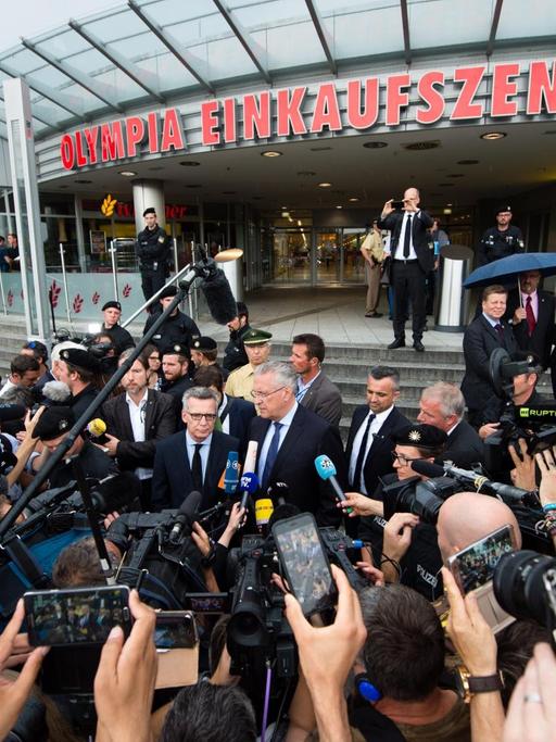 Bundesinnenminister Thomas de Maiziere (CDU, Mitte, l) und Bayerns Innenminister Joachim Herrmann (CSU, Mitte, r) geben einen Tag nach einer Schießerei mit Toten und Verletzten, vor dem Olympia-Einkaufszentrum (OEZ) in München (Bayern) ein Statement ab.