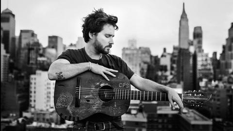 Ein Mann steht vor der Skyline einer amerikanischen Metropole, in der Hand hält er eine Gitarre.