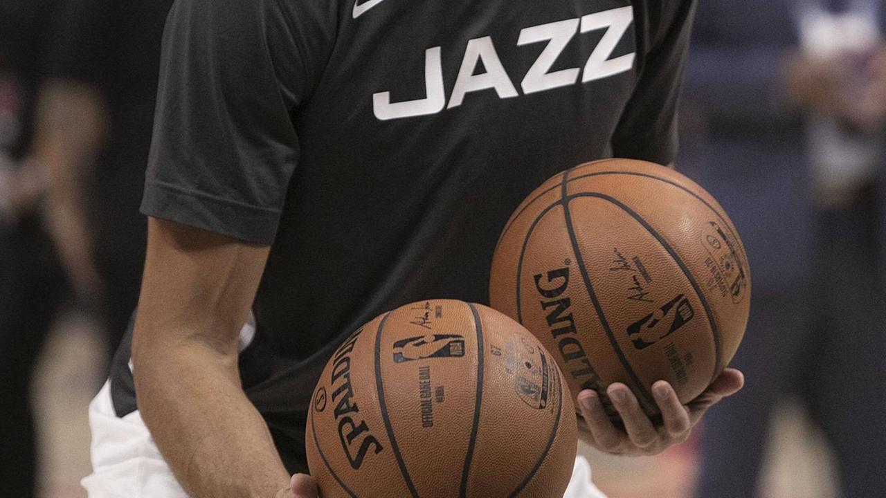 Auf dem Bild ist der Rumpf eines Mannes zu sehen, der ein schwarzes T-Shirt mit der weißen Aufschrift "Jazz" trägt und zwei Basketbälle in den Händen hält.