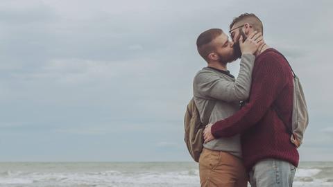 Zwei Männer küssen sich auf einem steinigen Strand, im Hintergrund ist das Meer zu sehen.