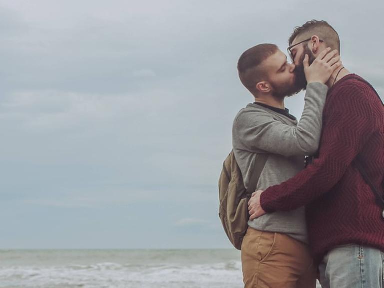 Zwei Männer küssen sich auf einem steinigen Strand, im Hintergrund ist das Meer zu sehen.