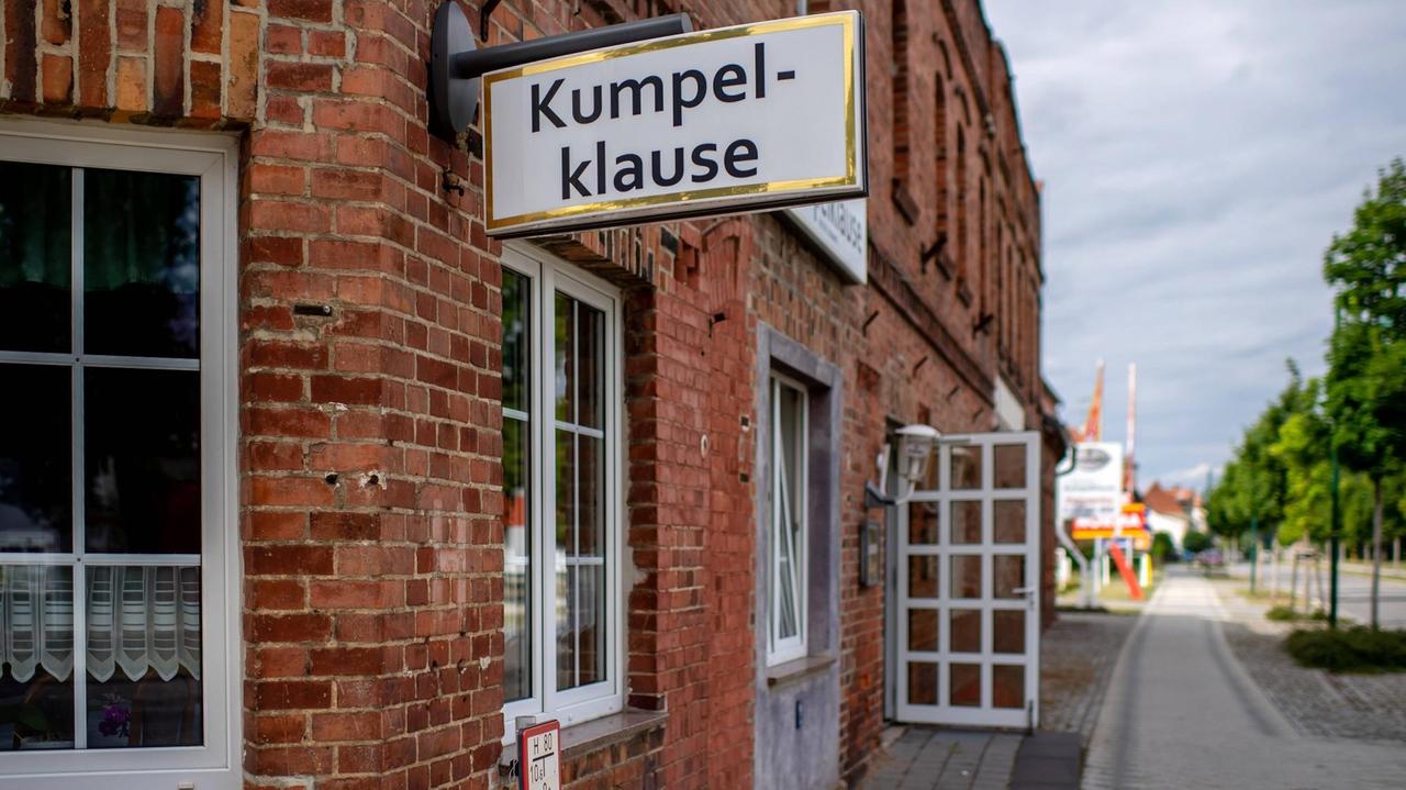 "Kumpel-Klause" steht an einem Haus aus Ziegelsteinen. Die Tür steht offen. Am rechten Bildrand verläuft die Dorfstraße in die Ferne.
