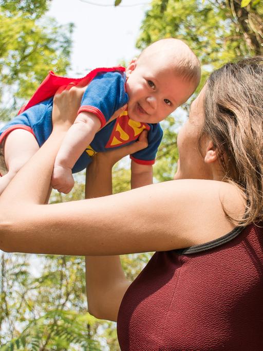 Eine Frau hebt ein Baby in einem Superman-Kostüm in die Luft.