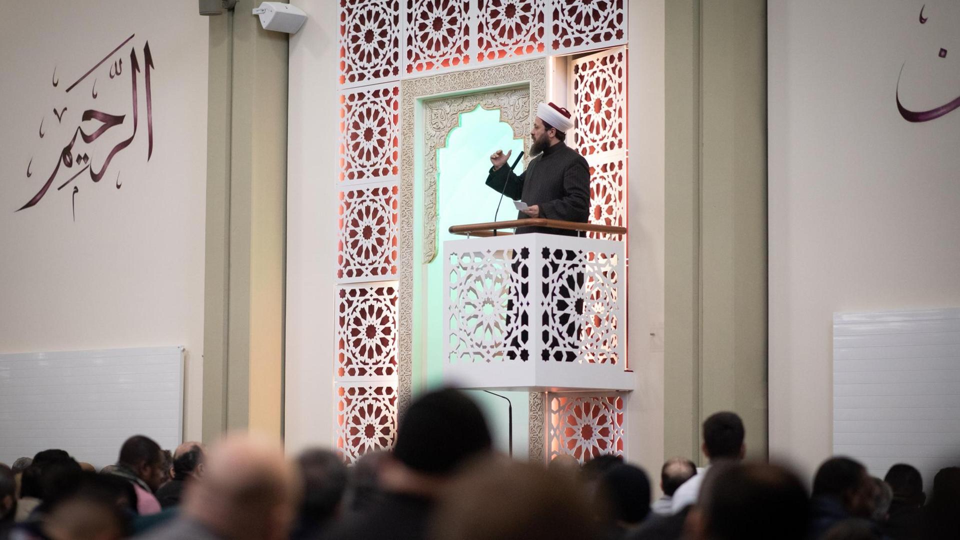 Ein Imam steht auf einem kleinen verzierten Balkon und spricht zu einer Gruppe Männer. An der Wand befinden sich arabische Schriftzeichen.