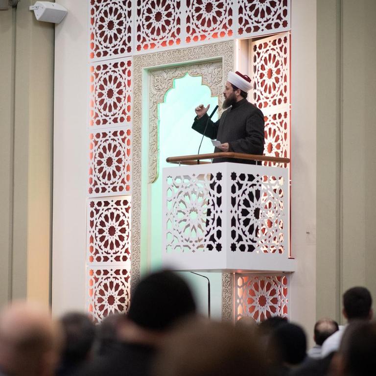 Ein Imam steht auf einem kleinen verzierten Balkon und spricht zu einer Gruppe Männer. An der Wand befinden sich arabische Schriftzeichen.