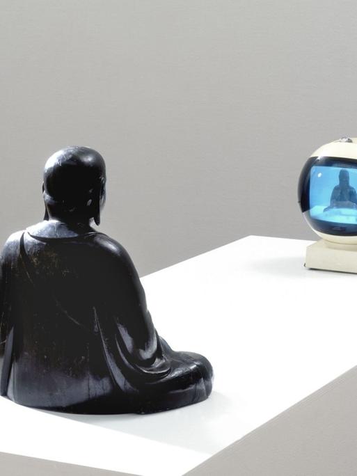 Die Installation TV-Buddha (1974) von Nam June Paik. - Eine Buddhafigur scheint sich selbst in einem Fernseher zu betrachten.