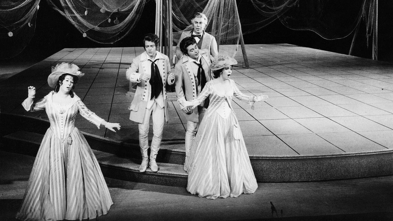 Auf einem Foto in schwarz-weiß stehen fünf Personen in historischen Kostümen auf einer Opernbühne.