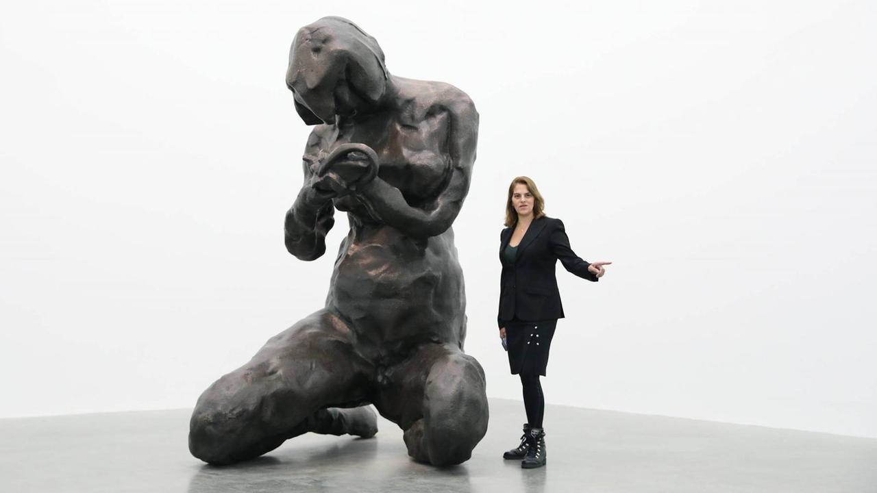 Ein weißer Raum, darin eine sehr große Skulptur, die mit groben Zügen eine menschliche Figur markiert, daneben etwa halb so groß die Künstlerin Tracey Emin.