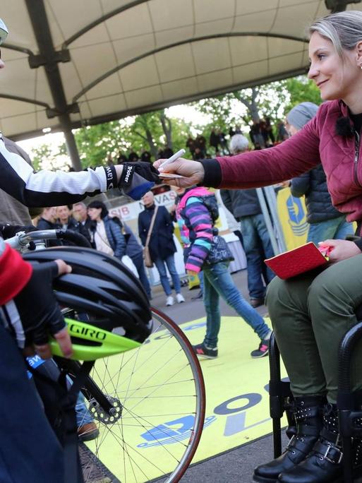 Kristina Vogel sitzt im Rollstuhl und übergibt einem Kind in Radkleidung ein Autogramm.