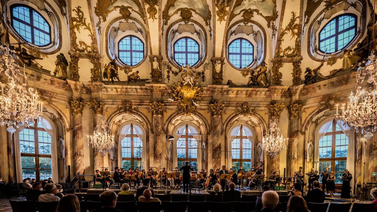 In einem hohen Saal mit vielen Fenstern und barocker Dekoration stehen Musiker und Sängerinnnen auf einer Bühne.