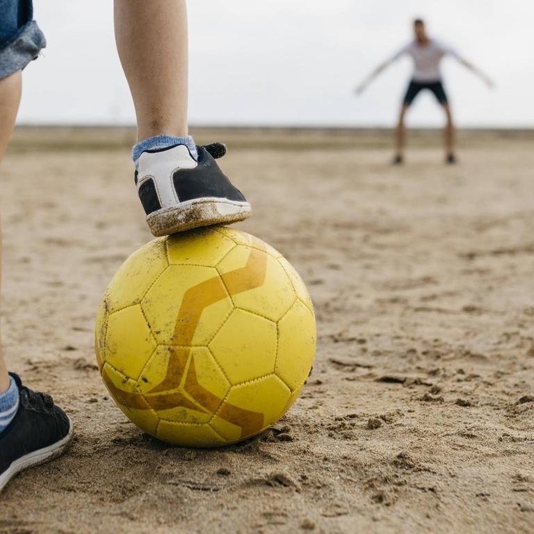 Die Beine eines Jungen mit Fußball, vor einem Tor mit Torwart