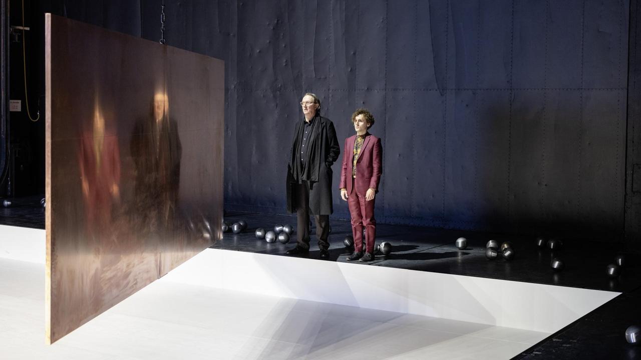 Ein älterer und ein junger Mann stehen am Rande einer Theaterbühne und schauen auf Abbilder ihrer selbst, die sich verzerrt von einer in der Mitte des Raumes hängenden Metallplatte widerspiegeln.