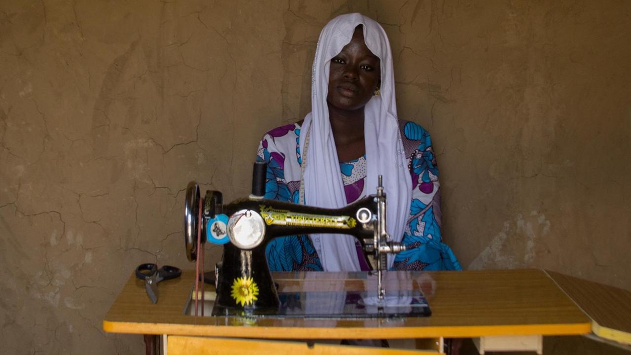 Eine junge afrikanische Frau mit weißem Kopftuch und buntem Kleid sitzt vor einer altmodischen Nähmaschine, die auf einem Klapptisch steht.