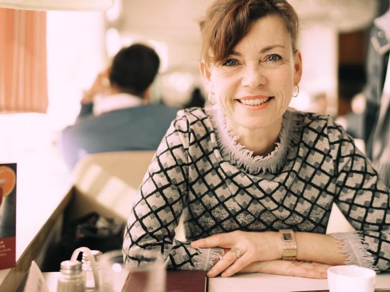 Renata Schmidtkunz sitzt in einem Cafe und lächelt in die Kamera.