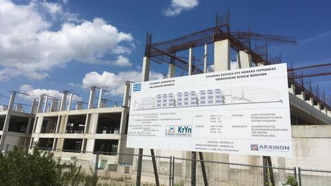 Das Bild zeigt den Rohbau der griechischen Schule in München. Vor dem Rohbau ist das Bauschild mit den Bauinformationen zu sehen.