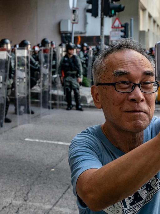 Ein älterer Mann macht ein Selfie von sich auf einer Straße in Hong Kong, mit Polizisten in voller Ausrüstung im Hintergrund, während der Demonstrationen gegen die Regierung.