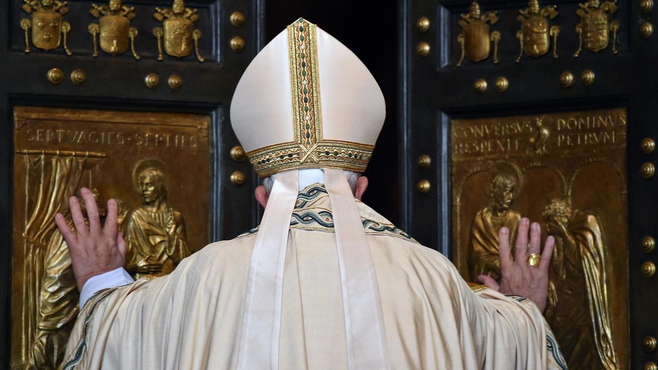 Man sieht den Papst von hinten, wie er die beiden Flügel der Pforte aufschiebt. Sie sind mit goldenen Ornamenten und Bildern verziert.