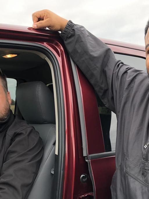 TM Garrett Schmid sitzt in seinem Auto, Ray Johnson lehnt von außen. Beide schauen in die Kamera.