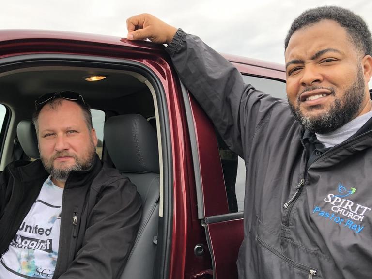 TM Garrett Schmid sitzt in seinem Auto, Ray Johnson lehnt von außen. Beide schauen in die Kamera.