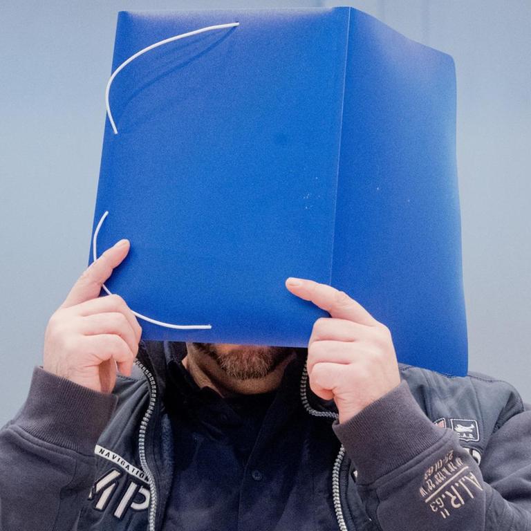 Der wegen vielfachen Mordes Angeklagte Niels Högel kommt in den Gerichtssaal und hält sich eine blaue Mappe vor das Gesicht