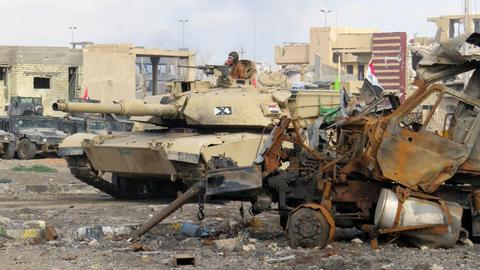 Zu sehen ist ein Panzer, dahinter zerschossene Häuser und im Vordergrund eine rostiges Autowrack.