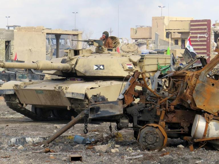 Zu sehen ist ein Panzer, dahinter zerschossene Häuser und im Vordergrund eine rostiges Autowrack.